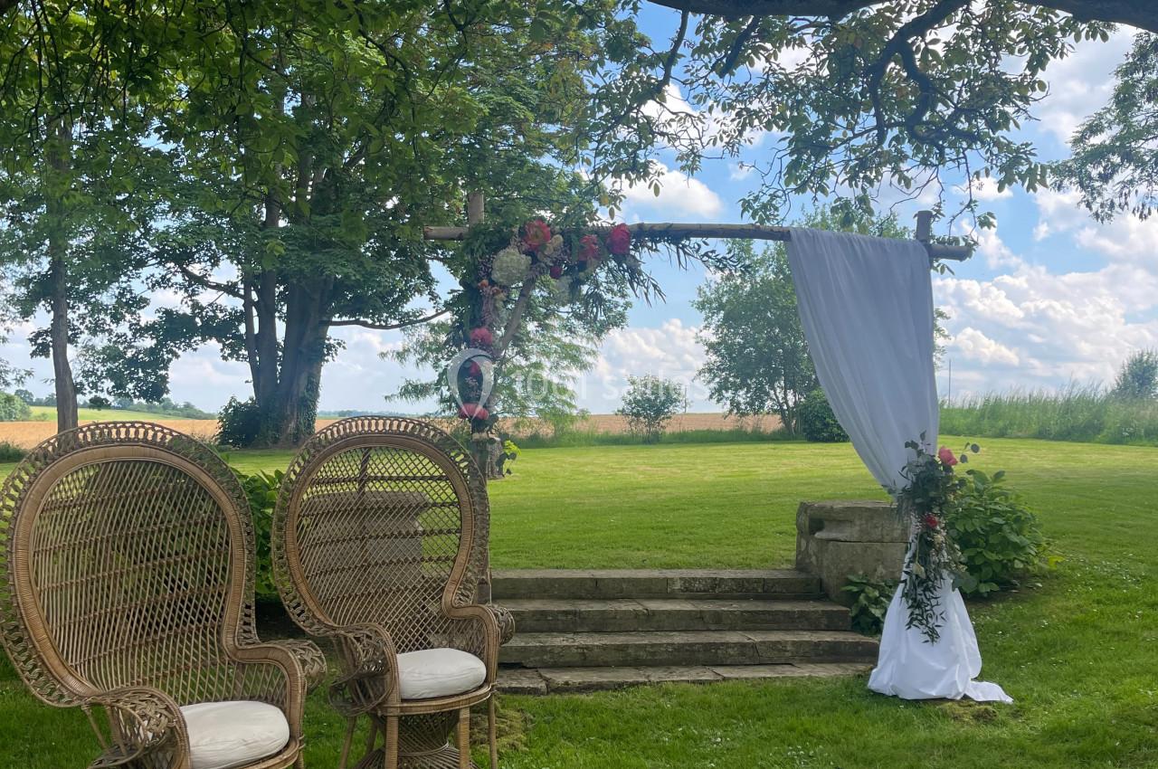 Deux fauteuils en rotin et une arche décorée de fleurs et de tissu blanc dans un jardin verdoyant.