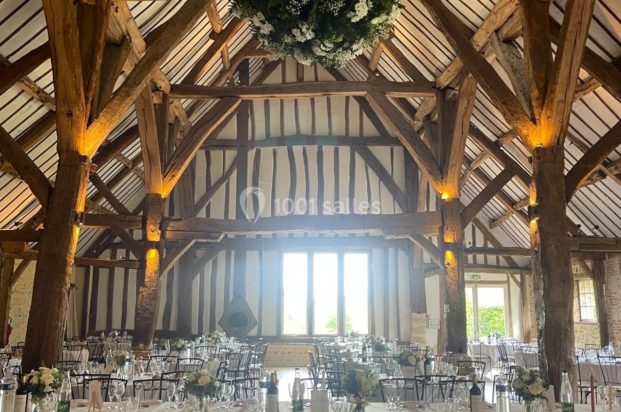 Grande salle de réception avec charpente en bois apparente, tables dressées et suspension florale centrale.