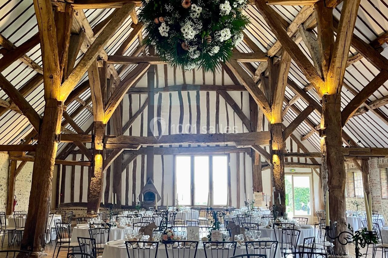 Salle de réception rustique avec poutres apparentes, tables dressées et une grande décoration florale suspendue.