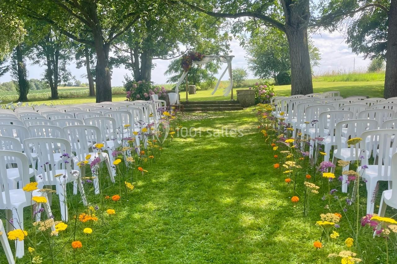 Allée centrale bordée de fleurs menant à une arche décorée pour une cérémonie en plein air, entourée d'arbres.