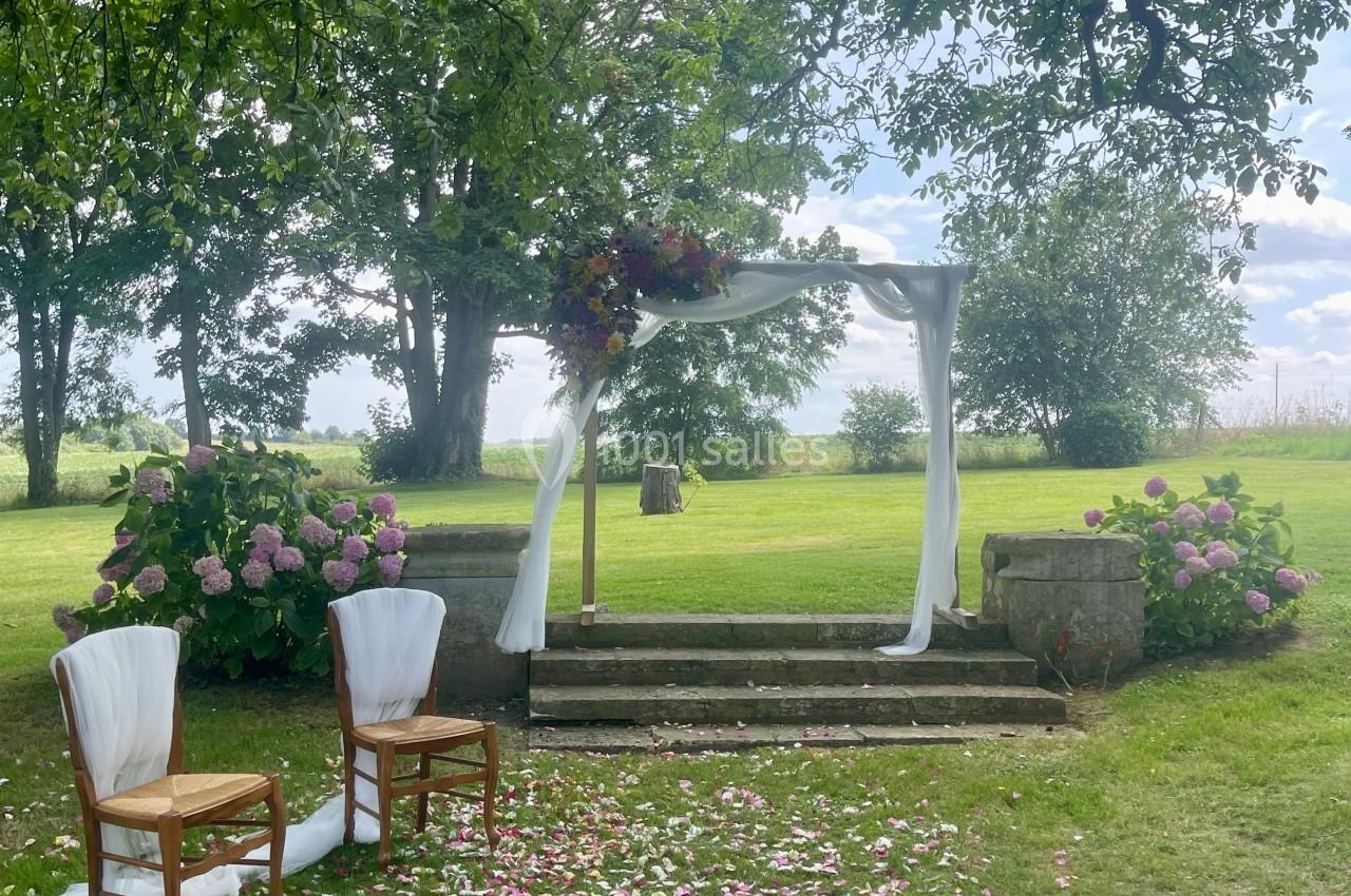 Arche de mariage décorée de fleurs et de voilages blancs dans un jardin verdoyant, avec des chaises et des pétales au sol.