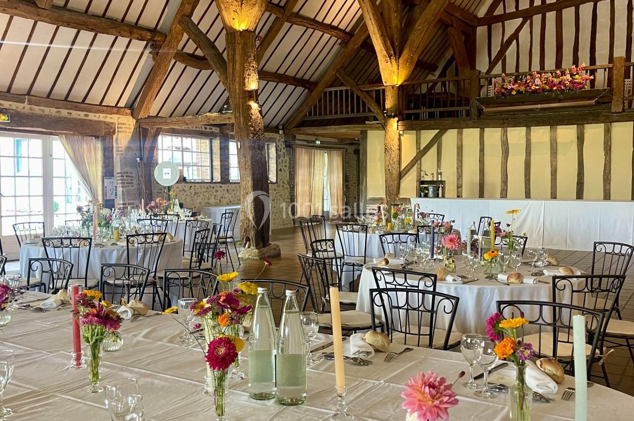 Salle de réception rustique avec poutres apparentes, tables dressées, décor floral coloré et lumière naturelle.
