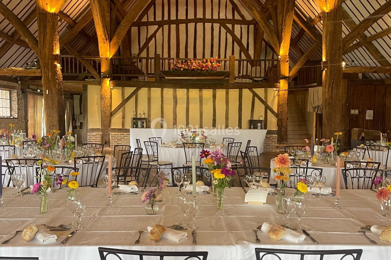 Salle de réception rustique avec poutres apparentes, tables dressées et décorations florales colorées.