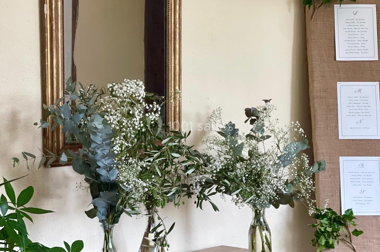 Trois vases en verre contenant des bouquets de feuillage et gypsophile posés sur une commode en bois devant un miroir.