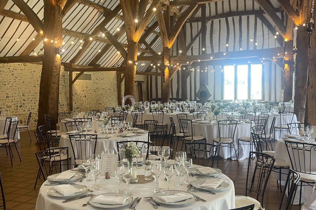 Salle de réception avec charpente en bois apparente, tables rondes dressées avec nappes blanches et chaises en métal.