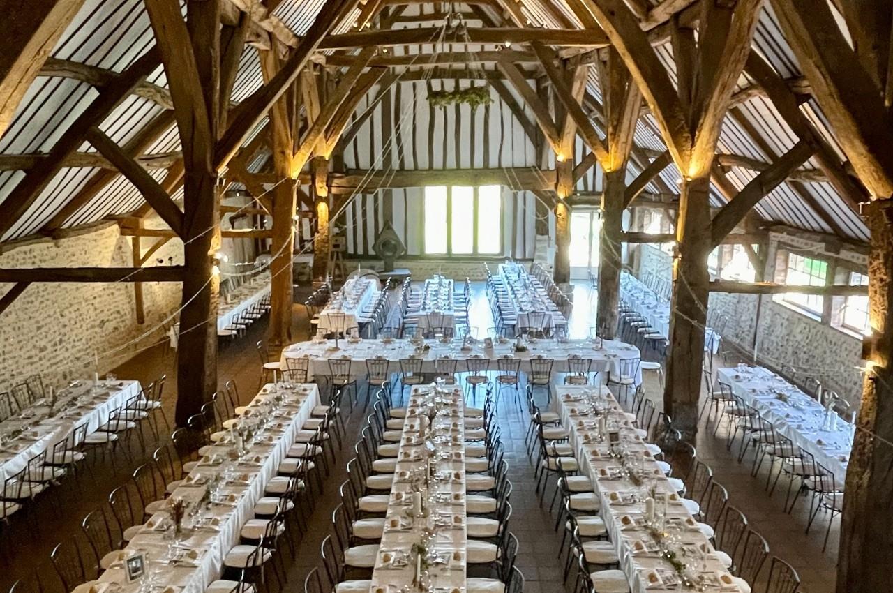 Salle de réception rustique avec de longues tables dressées pour un événement, sous une charpente en bois apparente.