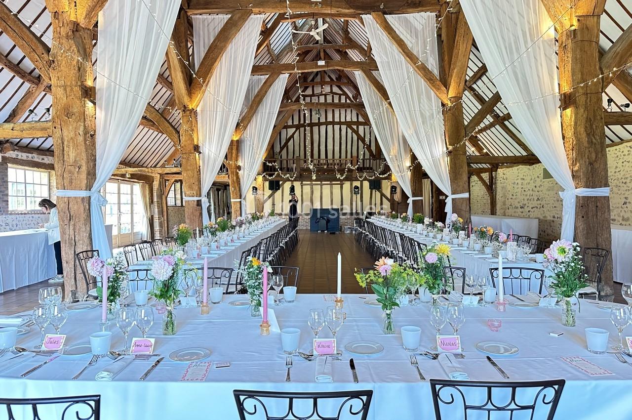Grande salle de réception rustique avec poutres apparentes, tables décorées de fleurs et vaisselle élégante.