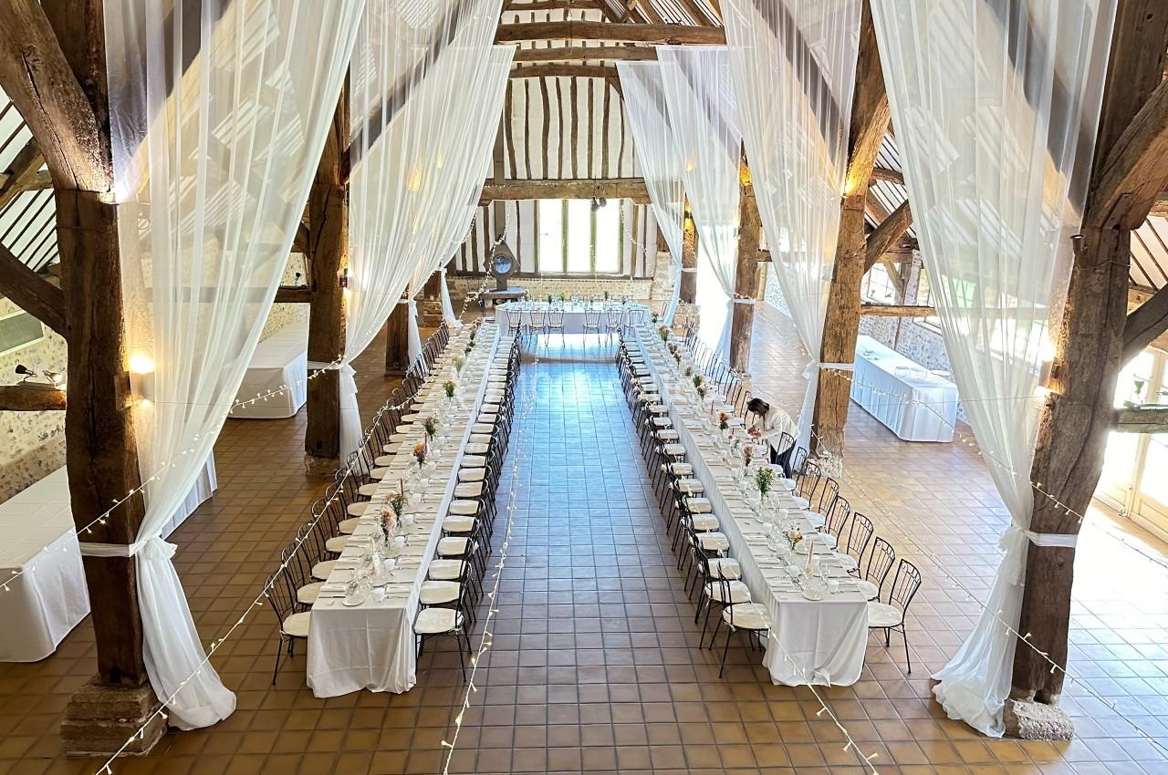 Grande salle de réception avec poutres apparentes, tables longues dressées et rideaux blancs suspendus.