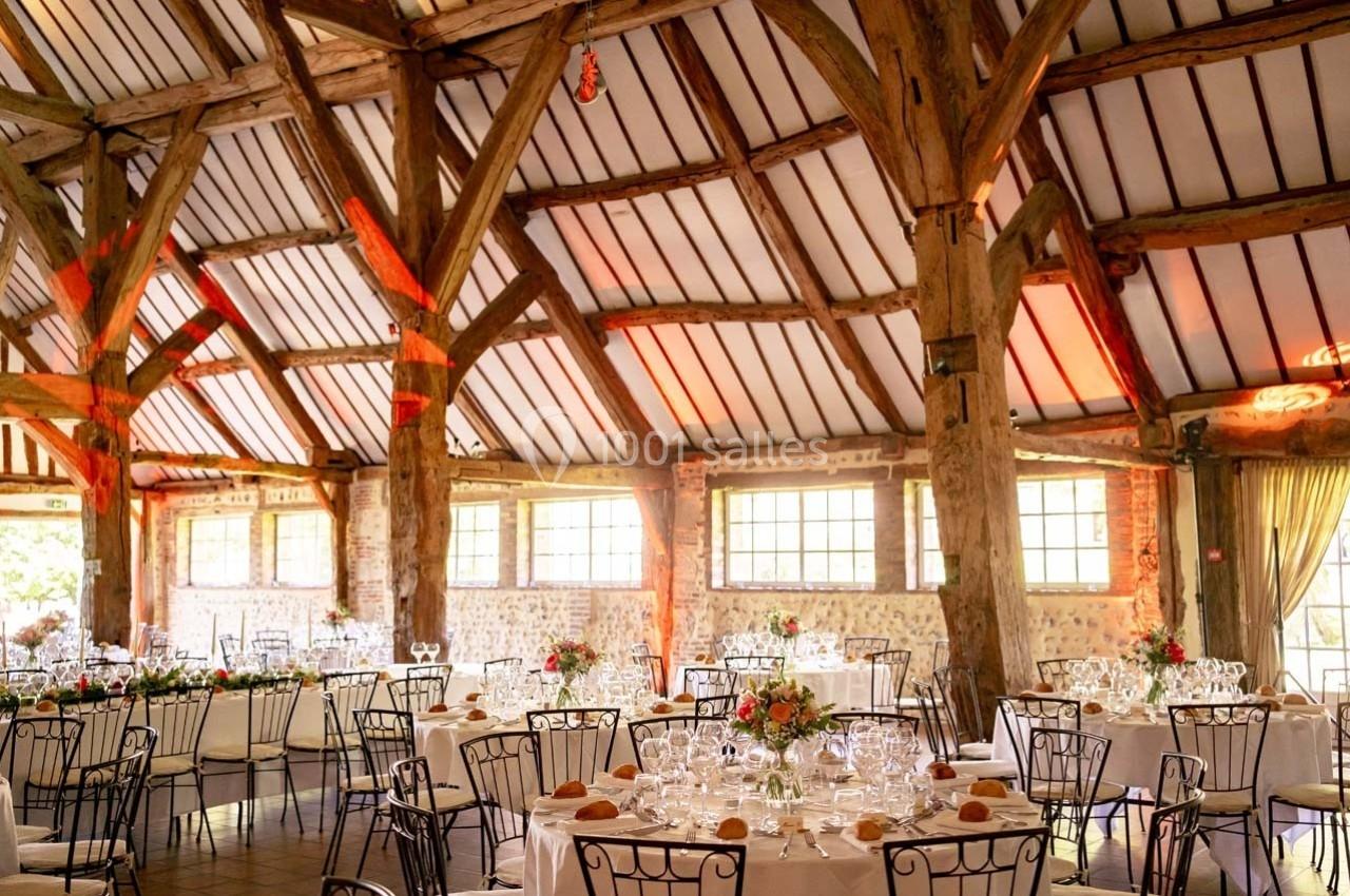 Salle de réception avec poutres apparentes, tables dressées avec nappes blanches, vaisselle et bouquets de fleurs.