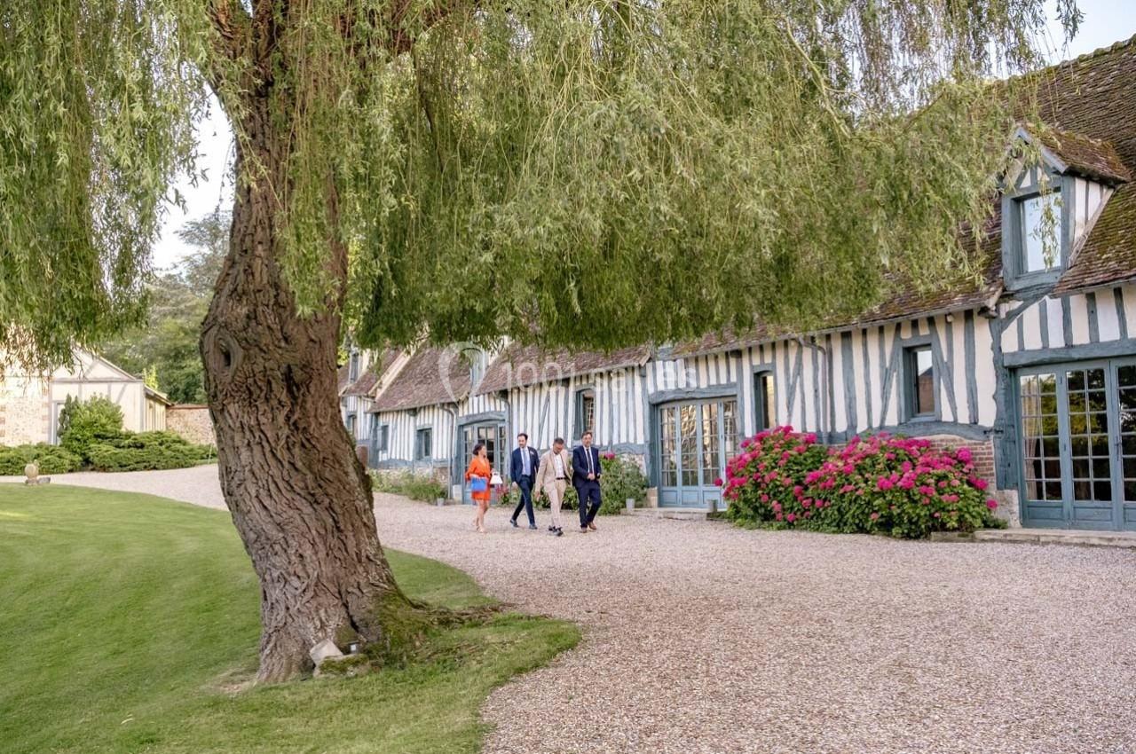 Maison à colombages avec toit en tuiles, entourée d'arbres, d'arbustes fleuris et d'un chemin en gravier.