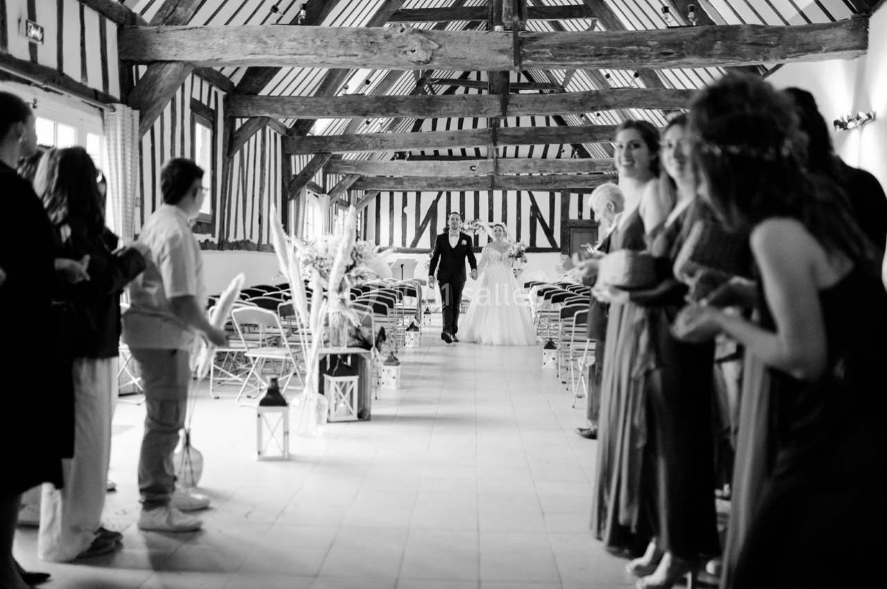 Un couple avance dans une allée centrale d'une salle en bois, entouré d'invités debout applaudissant.