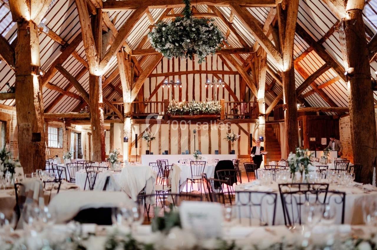 Salle de réception rustique avec poutres apparentes, tables décorées et un grand lustre floral suspendu.