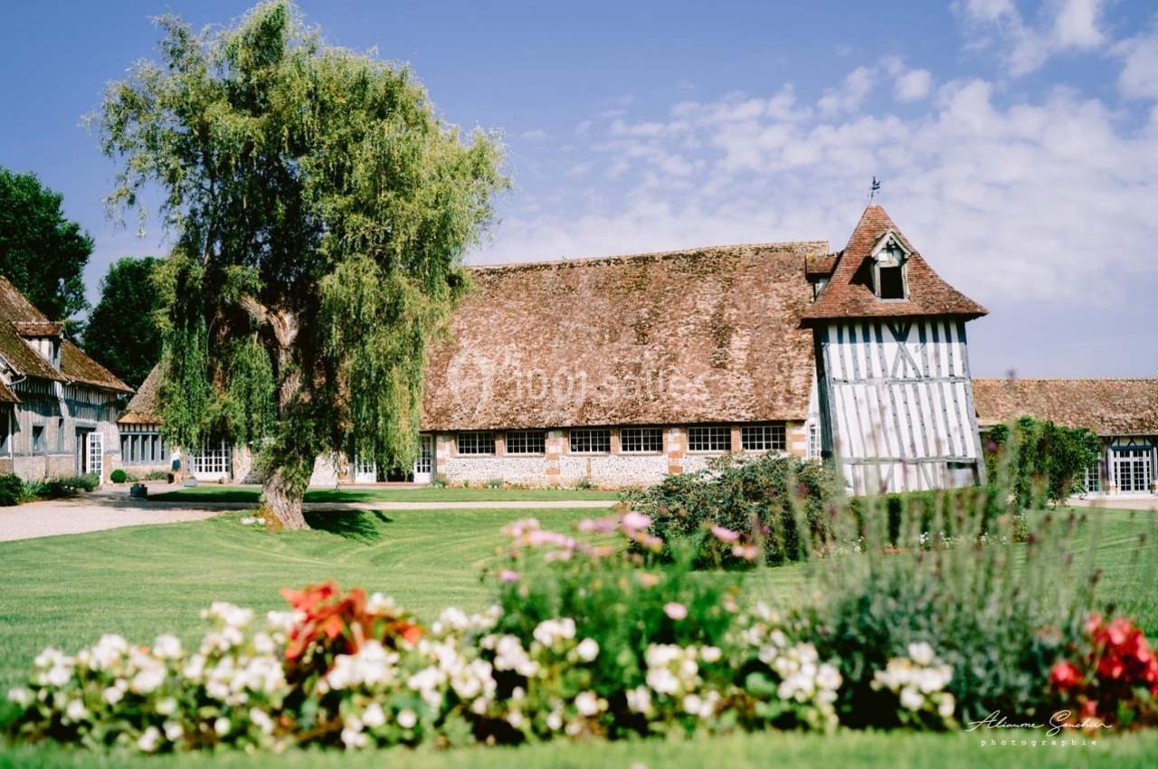Bâtiment à colombages avec toit en tuiles entouré d'un jardin fleuri et d'une pelouse sous un ciel bleu.