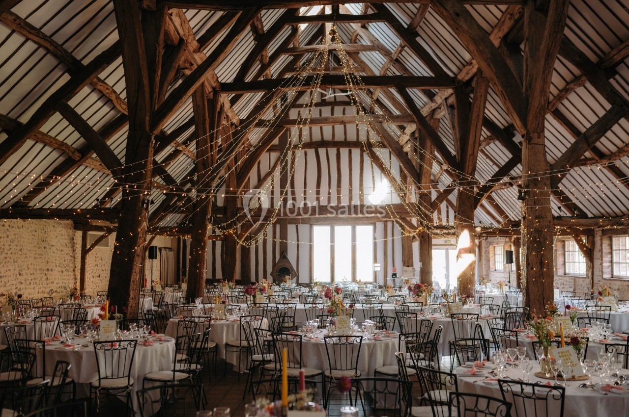 Salle de réception rustique avec poutres apparentes, tables rondes décorées et guirlandes lumineuses suspendues.