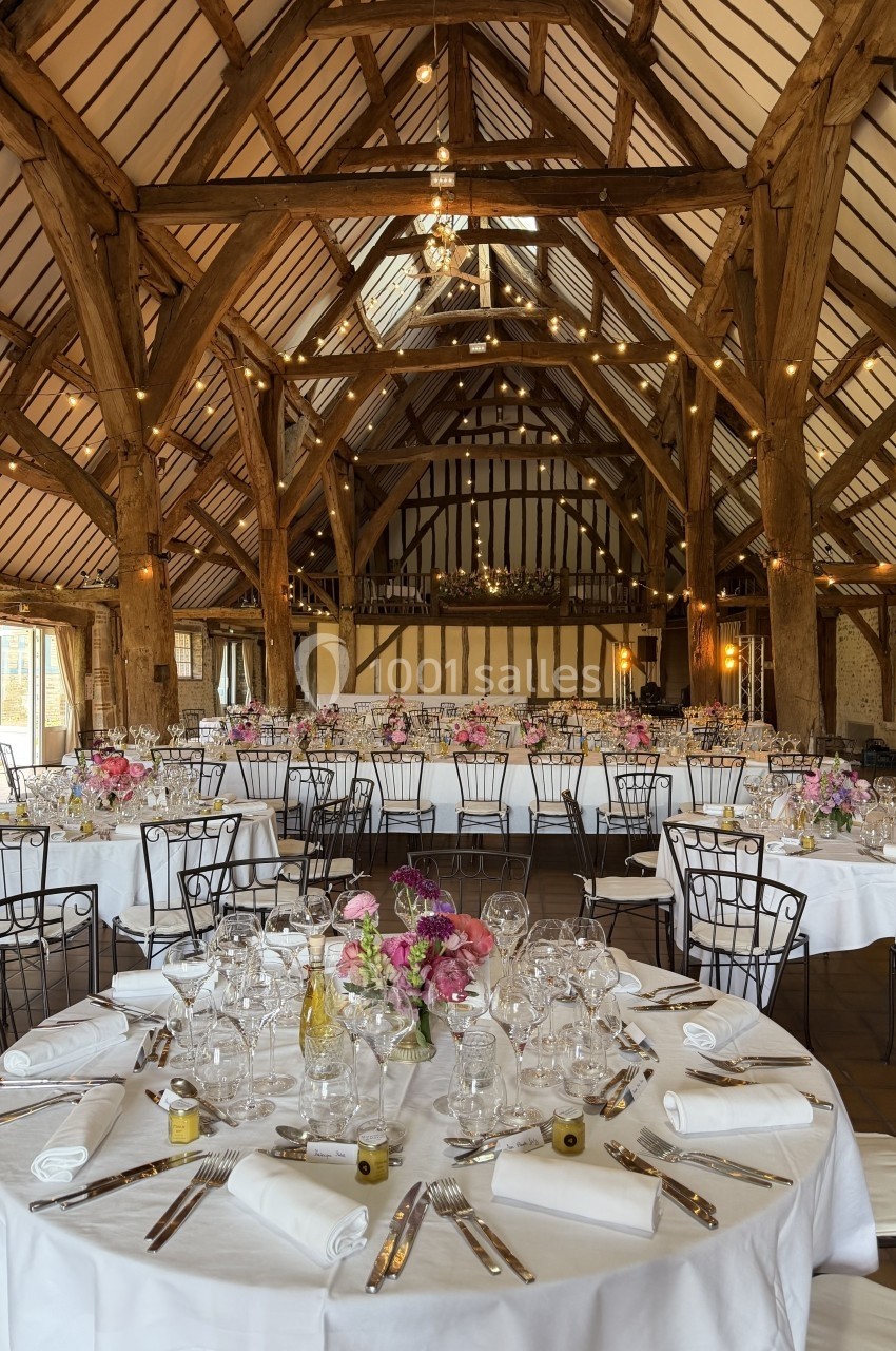 Grande salle de réception avec charpente en bois, tables rondes dressées avec nappes blanches et décorations florales.