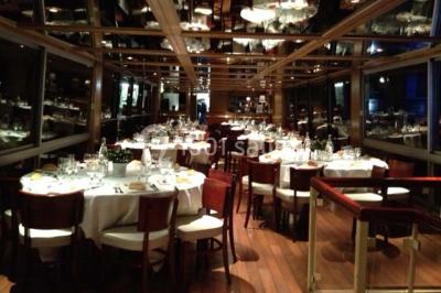 Salle de restaurant sur un bateau avec tables rondes dressées, nappes blanches et vue sur un paysage fluvial.