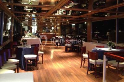 Salle de restaurant sur un bateau avec tables rondes dressées, nappes blanches et vue sur un paysage fluvial.
