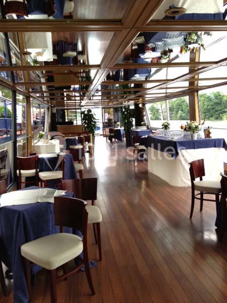 Salle de réception élégante avec tables dressées, nappes bleues et grandes baies vitrées donnant sur un paysage verdoyant.