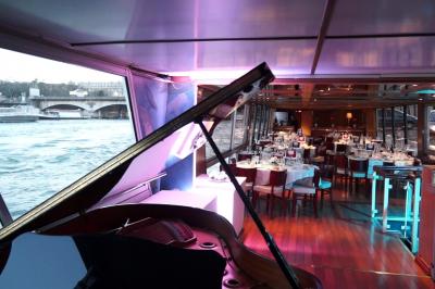 Salle de restaurant sur un bateau avec tables rondes dressées, nappes blanches et vue sur un paysage fluvial.