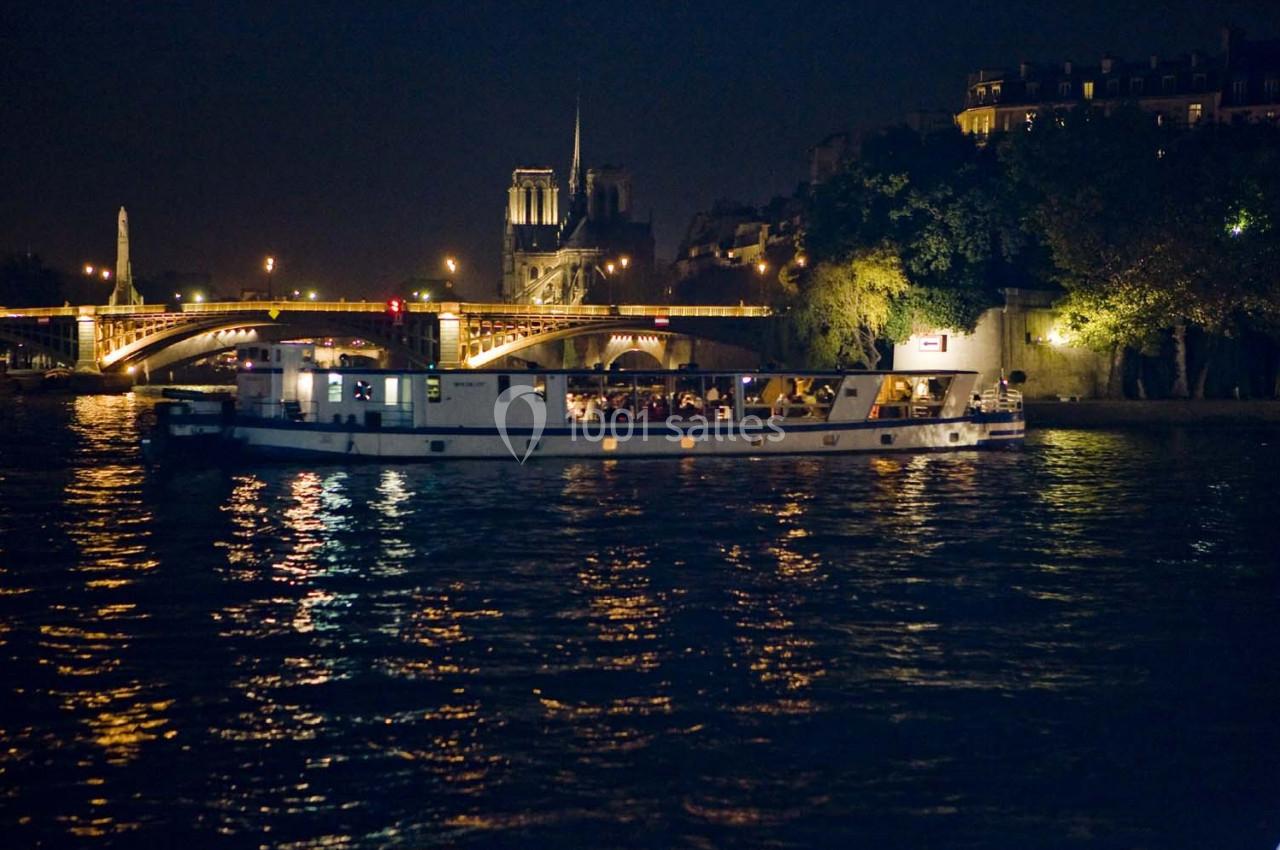 Bateau illuminé naviguant de nuit sur la Seine, avec Notre-Dame et un pont éclairé en arrière-plan.