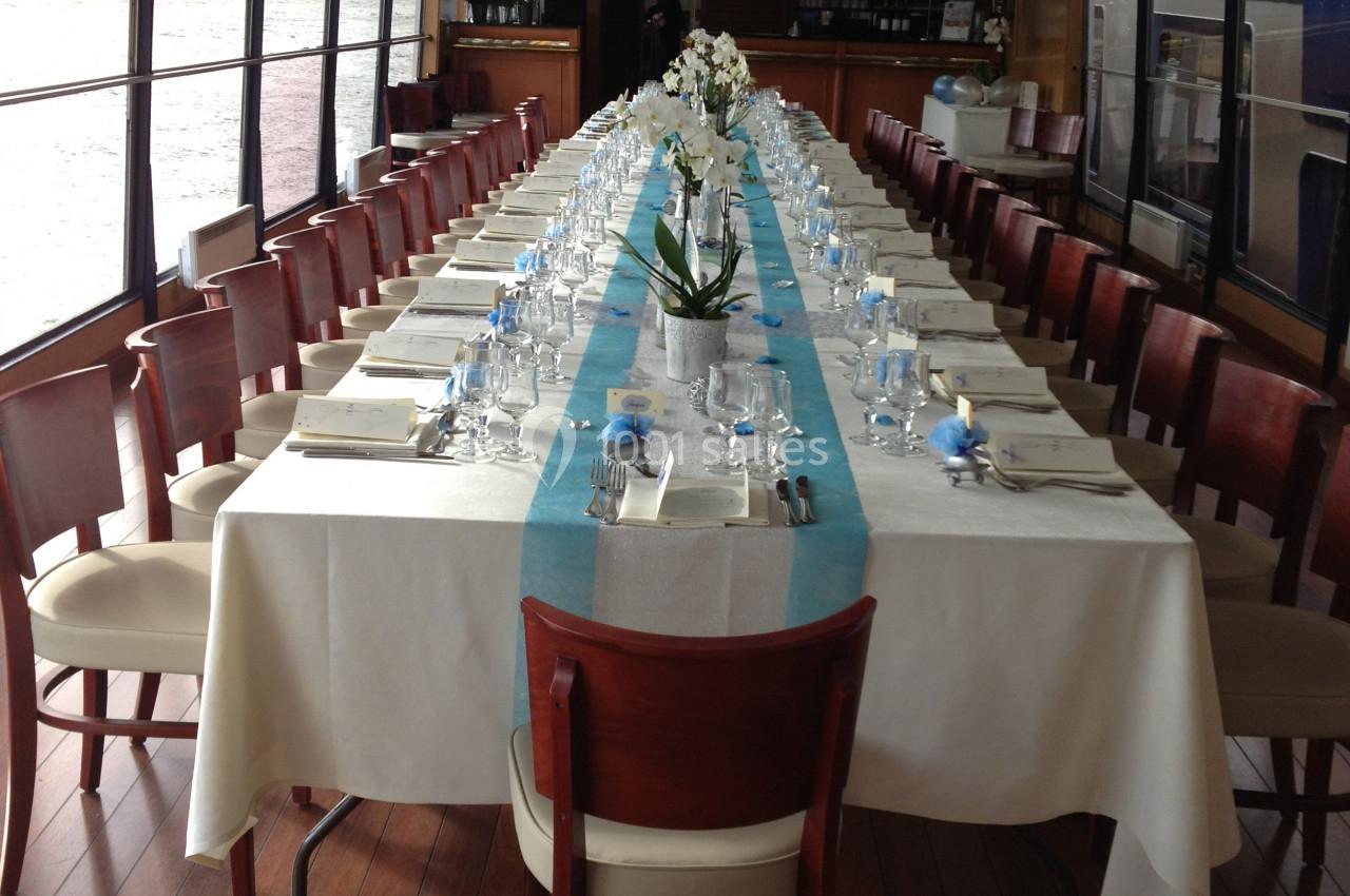 Grande table rectangulaire dressée avec nappes blanches, chemin de table bleu et orchidées, dans une salle vitrée.