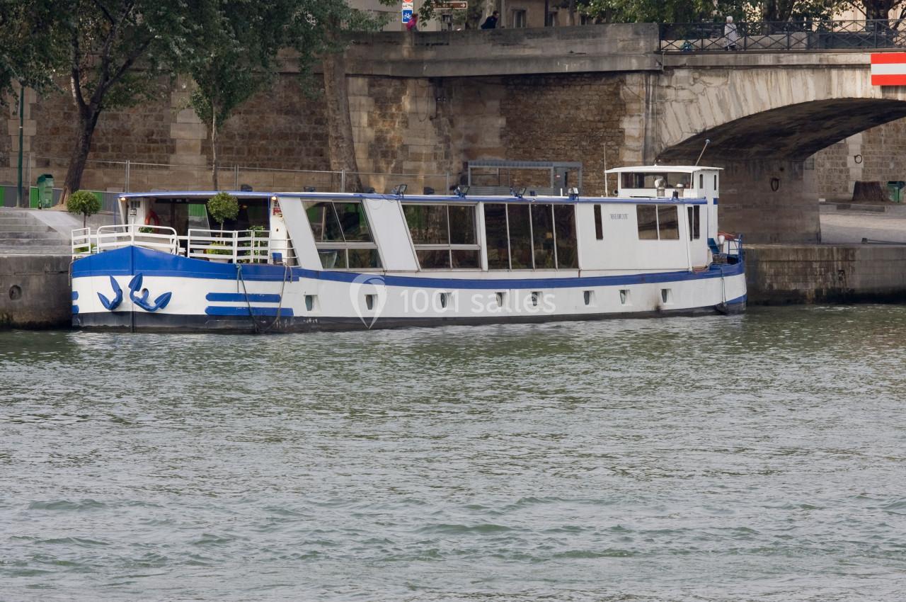 Bateau blanc et bleu amarré le long d'une rive avec un pont en pierre en arrière-plan.