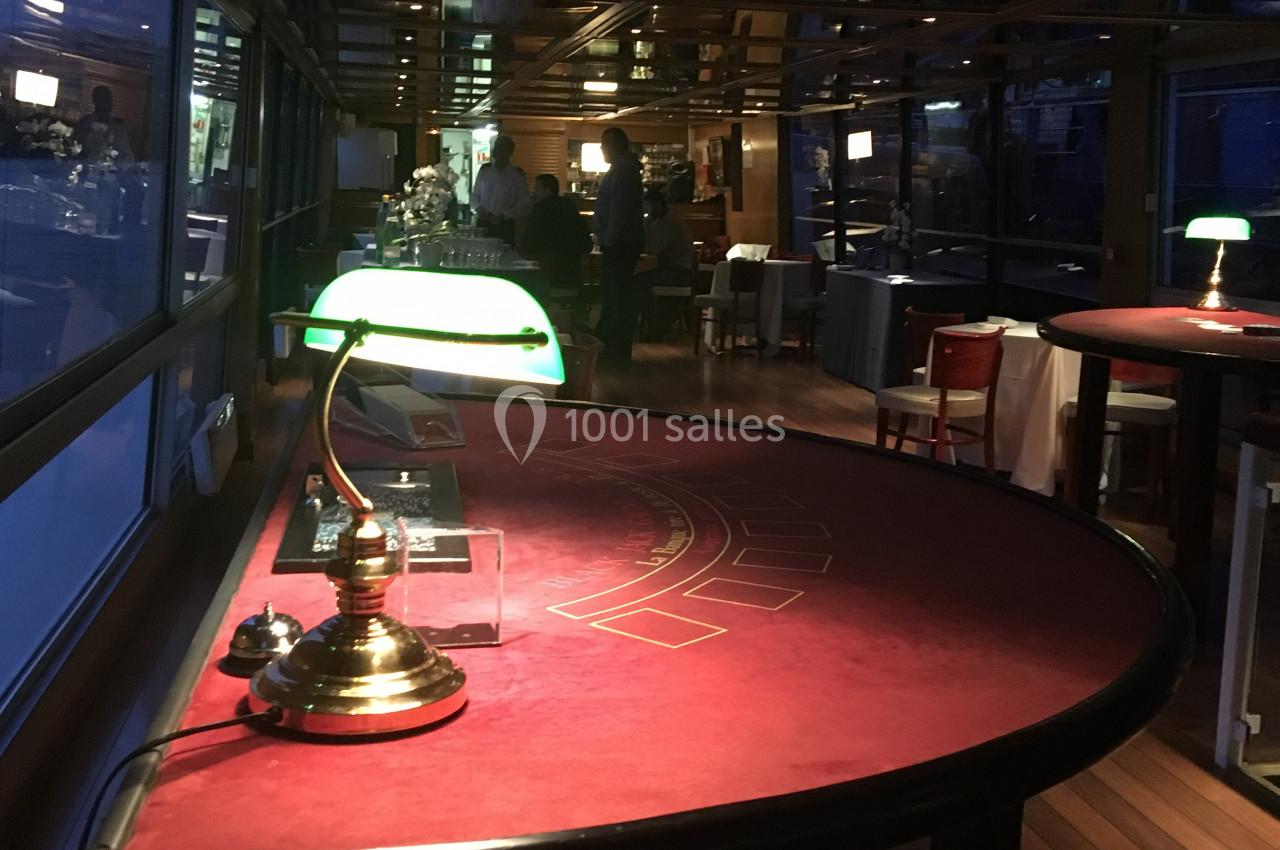 Table de jeu rouge éclairée par une lampe verte dans une salle sombre avec des fenêtres et des tables en arrière-plan.