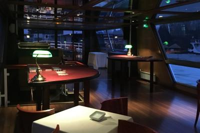 Salle de restaurant sur un bateau avec tables rondes dressées, nappes blanches et vue sur un paysage fluvial.