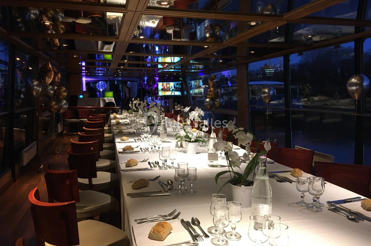 Salle de réception élégante avec une longue table dressée, décorée de fleurs blanches, vue sur l'extérieur nocturne.