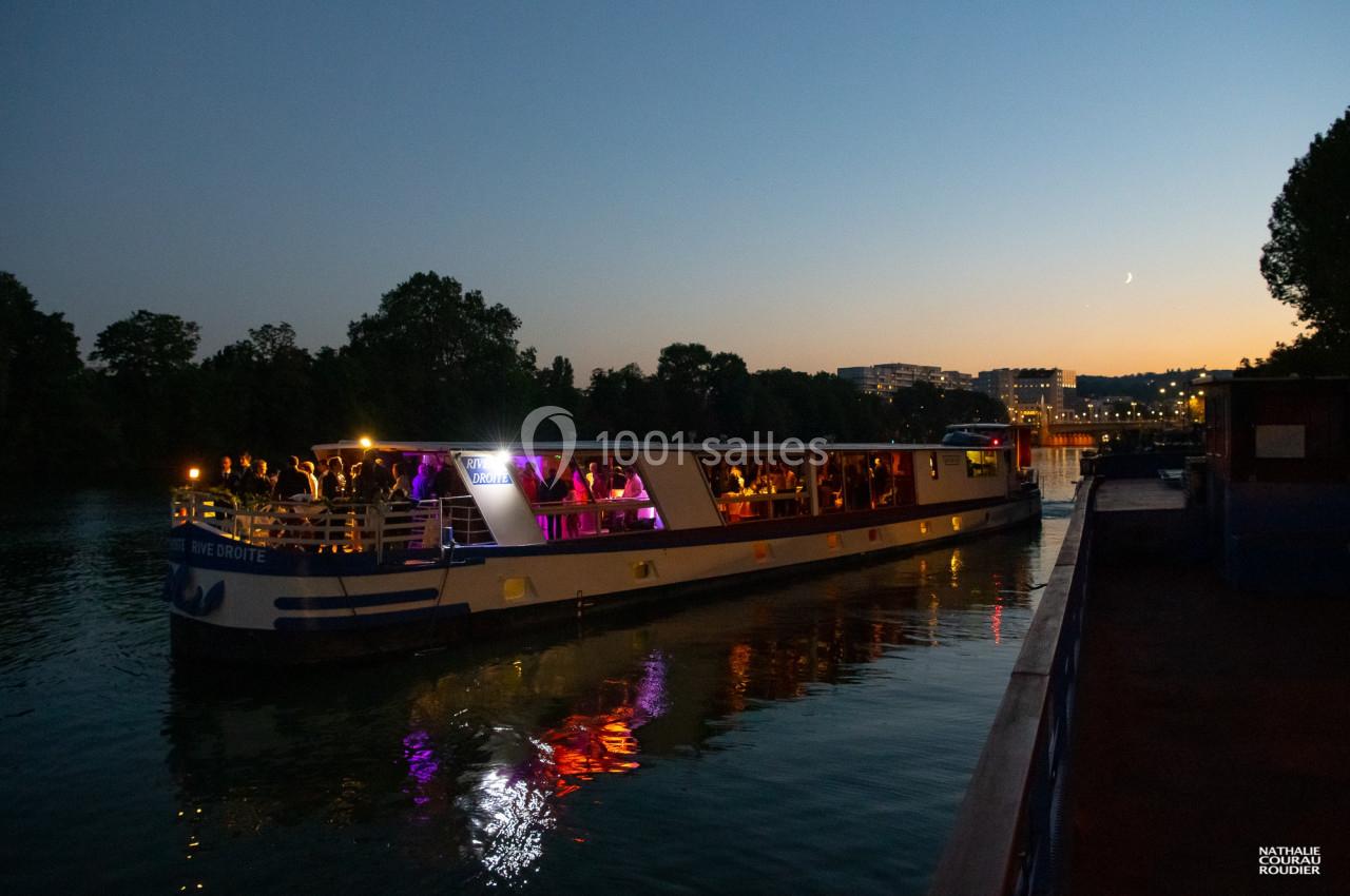 Bateau éclairé naviguant sur une rivière au crépuscule, avec des passagers visibles à bord.