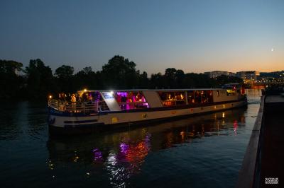Salle de restaurant sur un bateau avec tables rondes dressées, nappes blanches et vue sur un paysage fluvial.