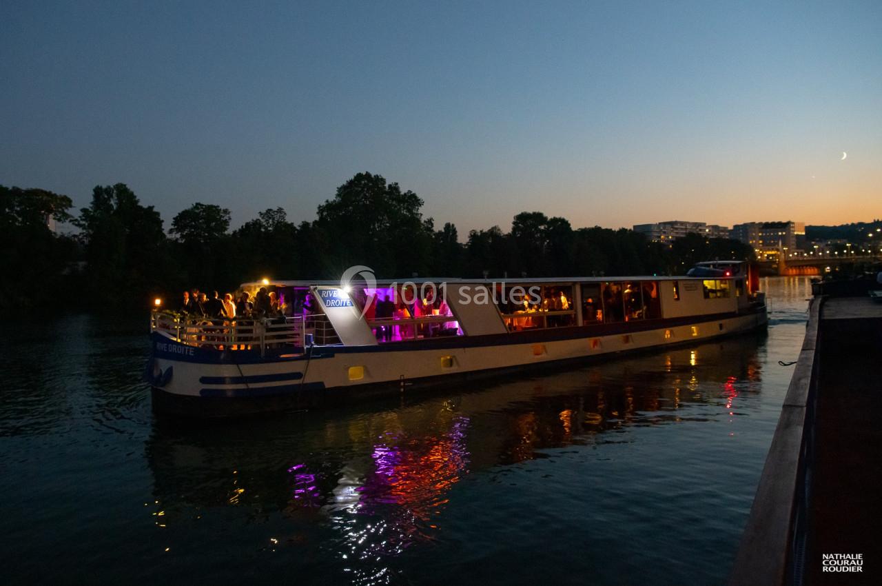 Bateau illuminé naviguant sur une rivière au crépuscule, avec des passagers visibles à bord.