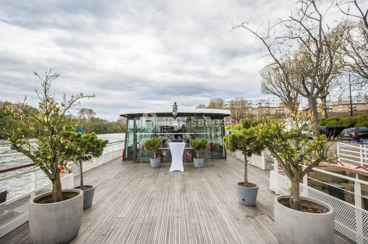 Terrasse en bois d'un bateau avec des plantes en pot, vue sur une salle vitrée et un fleuve en arrière-plan.