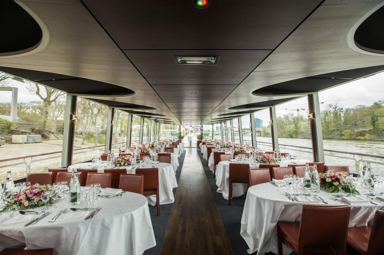 Salle de réception sur un bateau avec des tables rondes dressées, entourées de chaises en cuir, et vue sur un paysage…