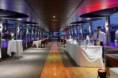 Salle de restaurant sur un bateau avec tables rondes dressées, nappes blanches et vue sur un paysage fluvial.