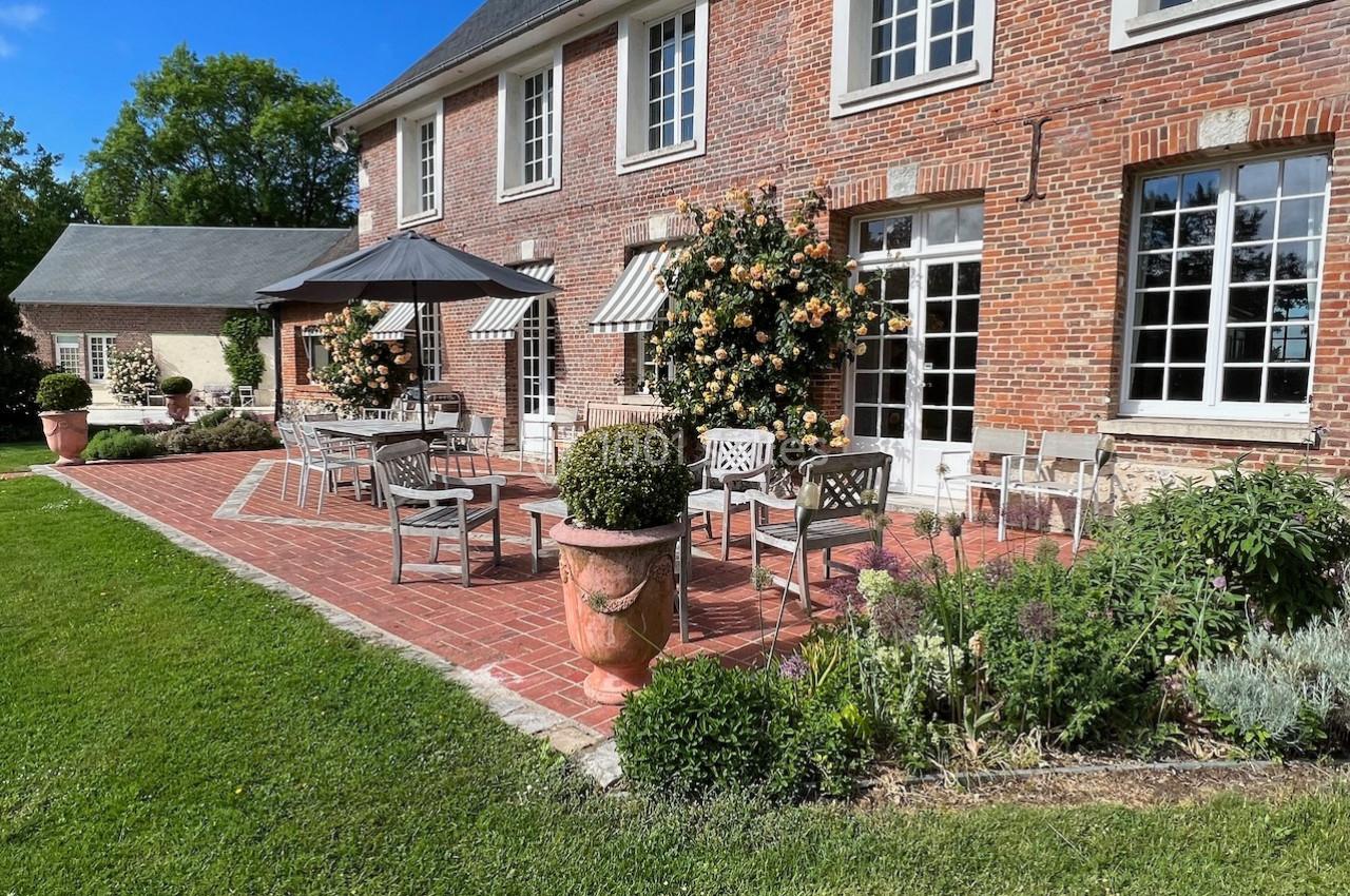 Terrasse en briques avec tables, chaises et parasols, bordée de fleurs et arbustes devant une maison en briques rouges.