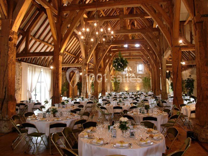 Salle de réception rustique avec poutres apparentes, tables rondes dressées et décorations lumineuses suspendues.