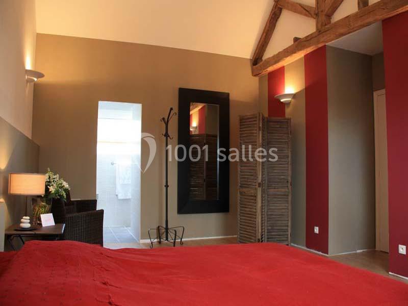 Chambre avec lit double, murs rouges et beiges, miroir mural, paravent en bois et accès à une salle de bain éclairée.