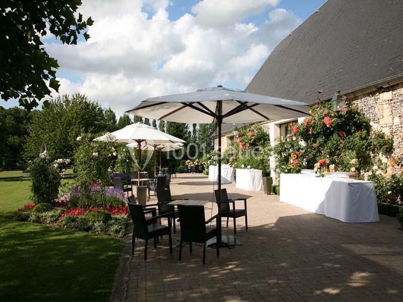 Terrasse pavée avec tables, chaises et parasols, entourée de fleurs et d'arbustes, près d'un bâtiment en pierre.