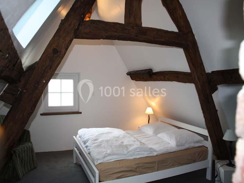 Chambre mansardée avec poutres apparentes, lit double, lampe de chevet allumée et fenêtre laissant entrer la lumière.