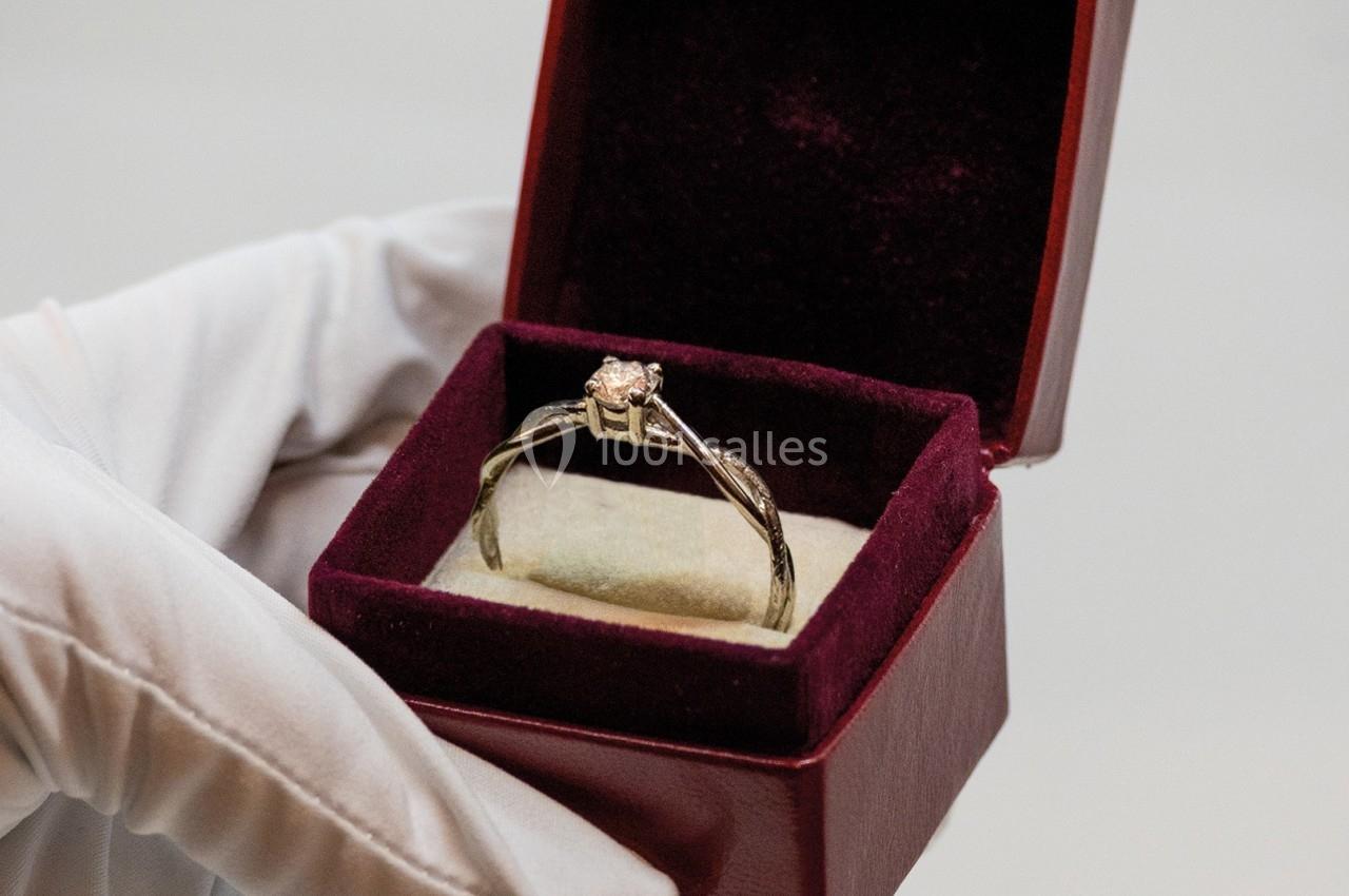 Une main gantée tient un écrin ouvert contenant une bague ornée d'une pierre précieuse.