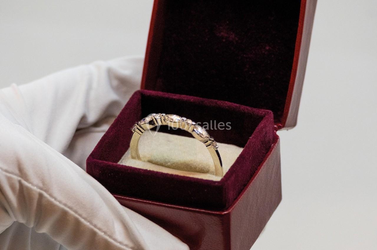 Une main gantée tient un écrin ouvert contenant une bague ornée de pierres brillantes.