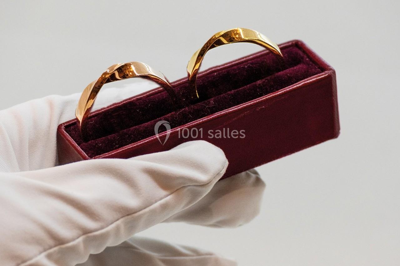 Deux alliances dorées dans un écrin rouge, tenues par une main gantée de blanc.