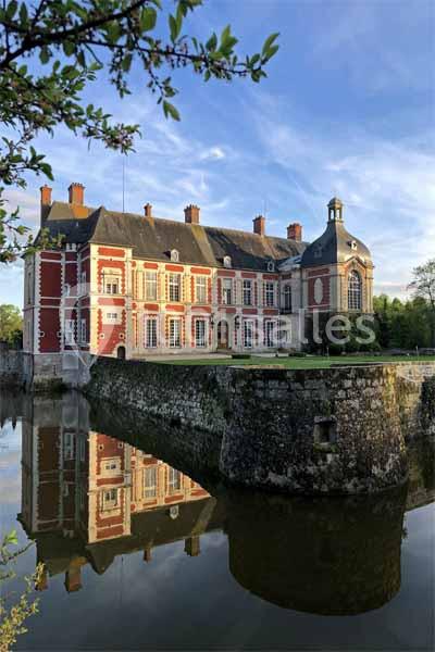 Façade d'un château en briques rouges et pierres blanches, entouré d'un fossé rempli d'eau, sous un ciel dégagé.