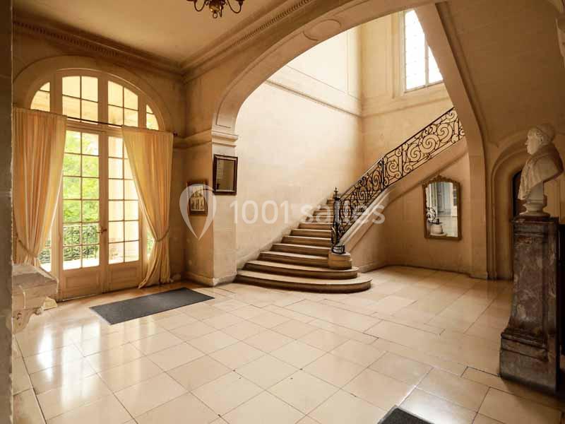 Hall d'entrée lumineux avec escalier en colimaçon, grandes fenêtres, rideaux beiges et statue sur un socle.