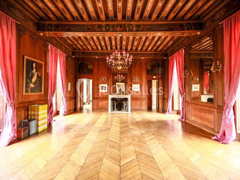 Salle avec parquet en chevrons, murs en bois, cheminée, lustres et rideaux roses encadrant de grandes fenêtres.