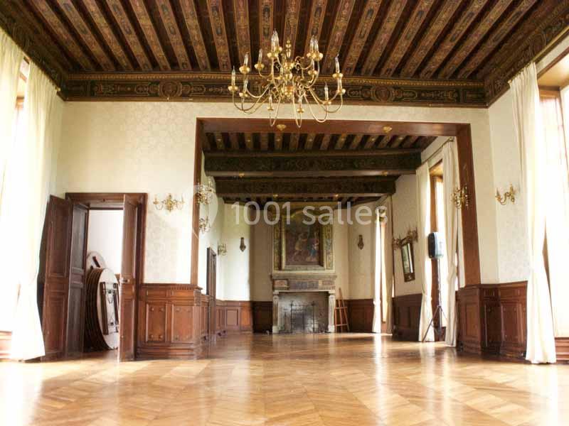 Salle spacieuse avec parquet en bois, poutres apparentes, lustres dorés et cheminée ornée d'un tableau.