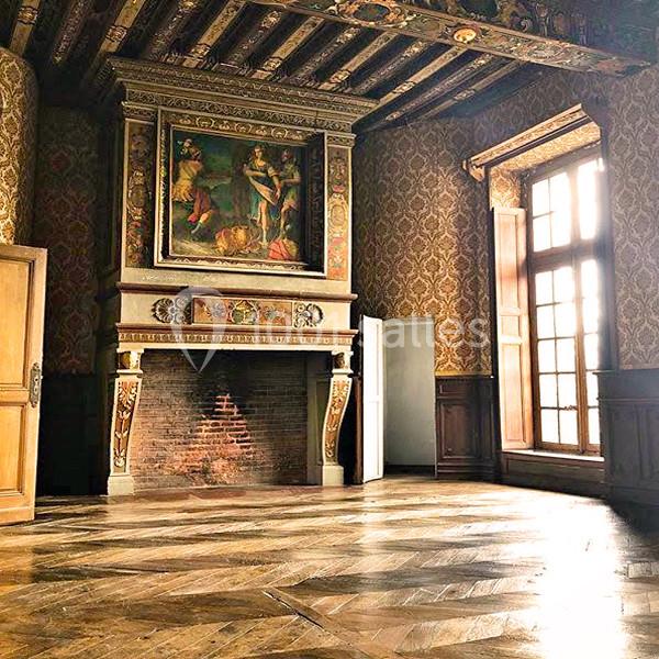 Salle ancienne avec cheminée ornée, tableau au-dessus, parquet en bois et grande fenêtre laissant entrer la lumière.