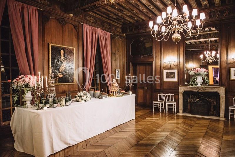 Salle élégante avec parquet, table de réception décorée, chandeliers, cheminée et portrait sur un mur boisé.