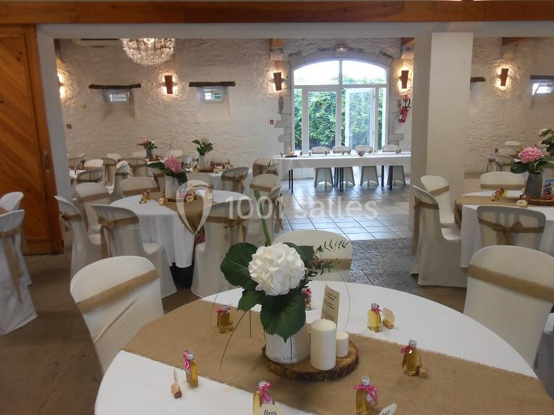 Salle de réception décorée avec des tables rondes, nappes blanches, centres de table fleuris et lumière naturelle.